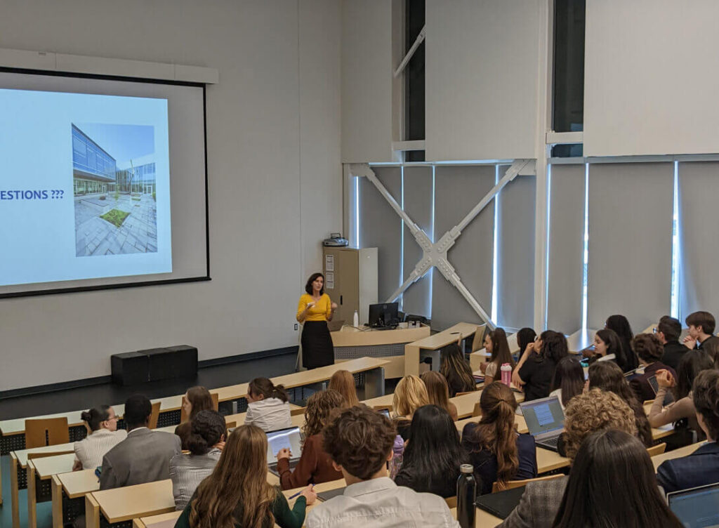 Conférencière présentant à un auditoire étudiant, salle lumineuse, écran de projection, interaction éducative, ambiance académique, étudiants attentifs, engagement et apprentissage.