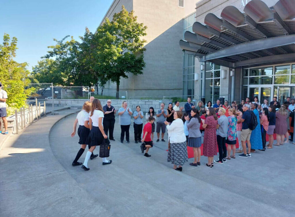 Étudiantes accueillies par des applaudissements, rassemblement devant le collège, ambiance ensoleillée, interaction chaleureuse, moment marquant de la rentrée scolaire.