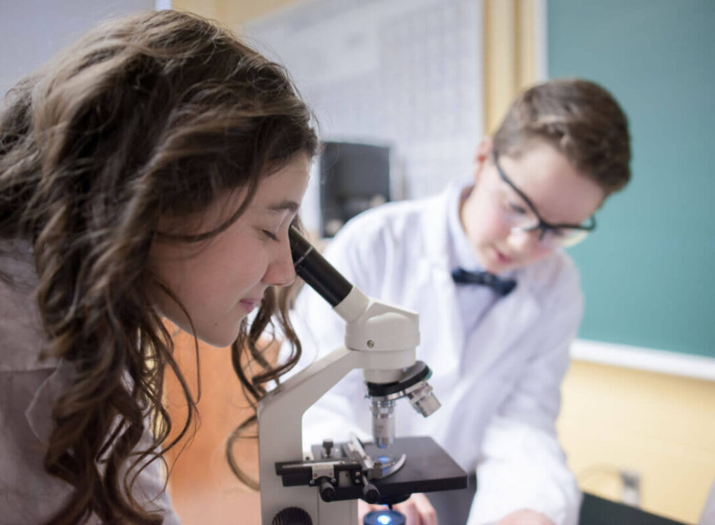 Étudiante regardant dans un microscope, camarade en arrière-plan, laboratoire de sciences, blouse blanche, apprentissage pratique, salle de classe, matériel scientifique, environnement scolaire, exploration et curiosité scientifique.