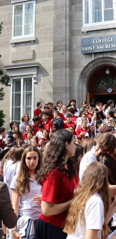 Groupe d'étudiants rassemblés devant l'entrée du Collège Saint-Sacrament, bâtiment historique en pierre grise, ambiance animée, uniformes rouges et blancs, journée scolaire, rassemblement, moment communautaire, esprit de camaraderie, tradition académique.