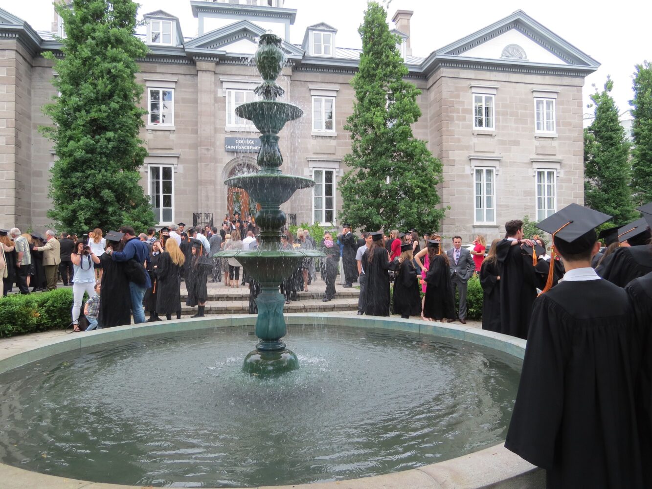 Cérémonie de remise des diplômes devant une fontaine, étudiants en toge noire, bâtiment historique du Collège Saint-Sacrament, familles réunies, célébration académique, Fondation Saint-Sacrament, événement officiel, soutien des partenaires et donateurs.