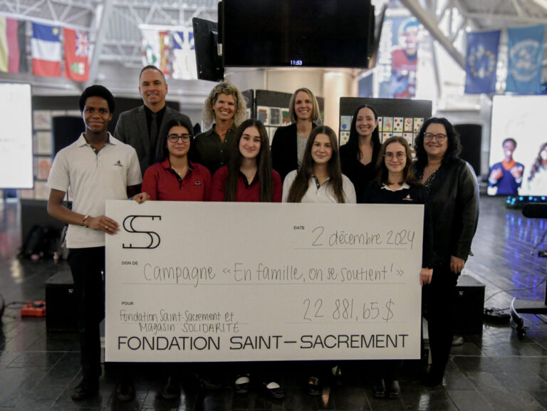 Photo de groupe, campagne "En famille, on se soutient!", grand chèque de 22 881,65 $, élèves et membres de la Fondation Saint-Sacrament, collaboration avec Magasin Solidarité, ambiance solidaire, décor intérieur, drapeaux internationaux en arrière-plan.