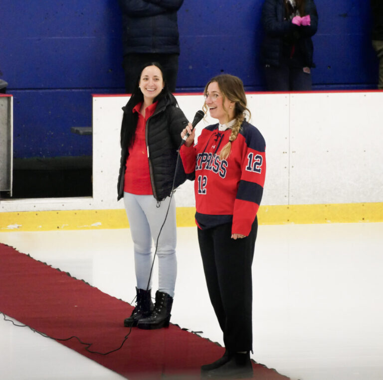 Deux femmes, discours sur la glace, l'une portant un chandail de hockey rouge "Express 12", ambiance sportive, tapis rouge, patinoire, engagement communautaire, Fondation Saint-Sacrament, Collège Saint-Sacrament, spectateurs en arrière-plan.