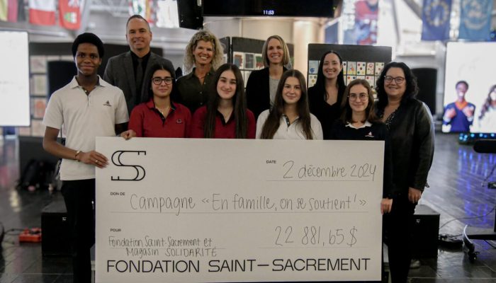 Photo de groupe, campagne "En famille, on se soutient!", grand chèque de 22 881,65 $, élèves et membres de la Fondation Saint-Sacrament, collaboration avec Magasin Solidarité, ambiance solidaire, décor intérieur, drapeaux internationaux en arrière-plan.