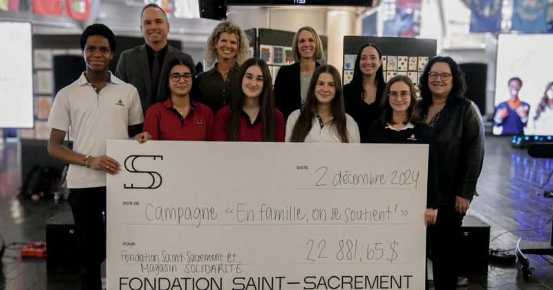 Photo de groupe, campagne "En famille, on se soutient!", grand chèque de 22 881,65 $, élèves et membres de la Fondation Saint-Sacrament, collaboration avec Magasin Solidarité, ambiance solidaire, décor intérieur, drapeaux internationaux en arrière-plan.