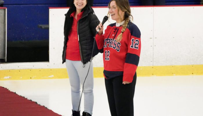 Deux femmes, discours sur la glace, l'une portant un chandail de hockey rouge "Express 12", ambiance sportive, tapis rouge, patinoire, engagement communautaire, Fondation Saint-Sacrament, Collège Saint-Sacrament, spectateurs en arrière-plan.